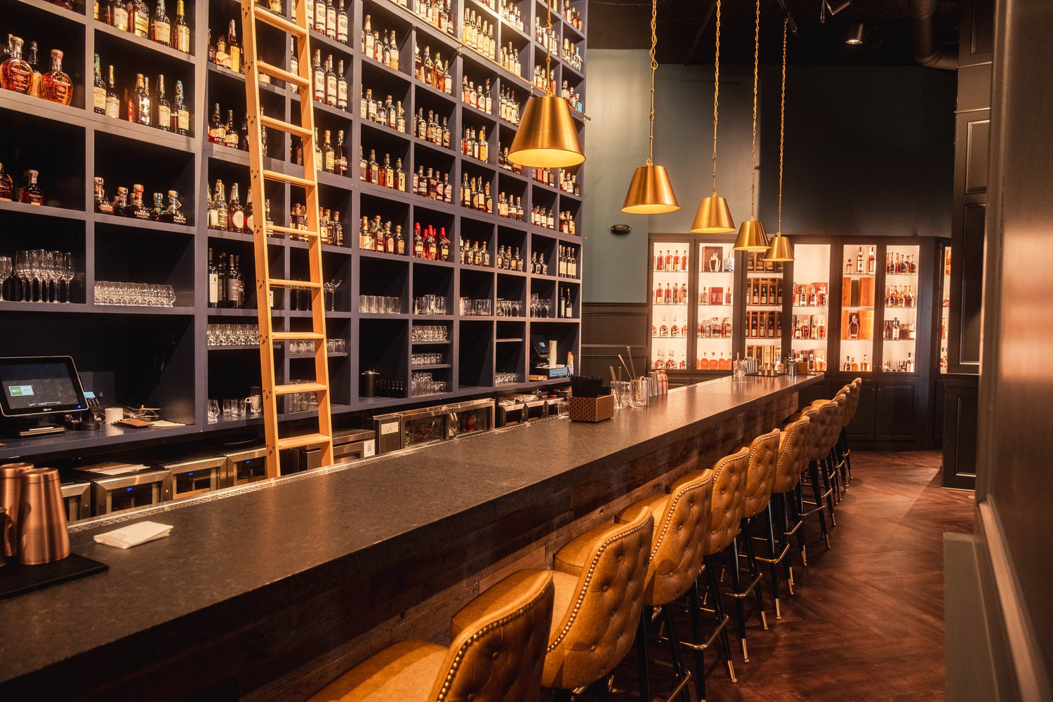 Bar with shelves stocked with bottles, a long bar counter, and gold pendant lights.